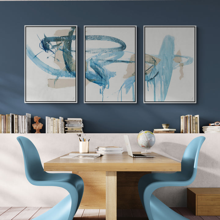 A modern workspace featuring three blue canvas triptych art, a wooden table, blue chairs, and a bookshelf against a navy wall.