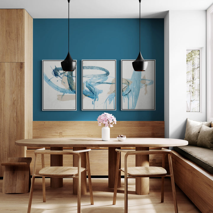 A modern dining area features a round wooden table, three canvas triptych art paintings on a teal wall.