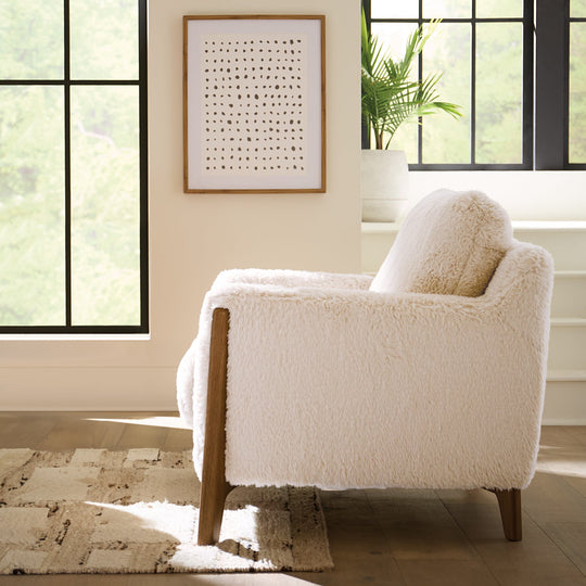 Side view of a cream-colored faux-shearling accent chair with mid-century modern wooden legs in bedroom.