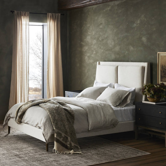 Cozy bedroom with a light-colored bed, plush pillows, and soft bedding, illuminated by natural light through sheer curtains.