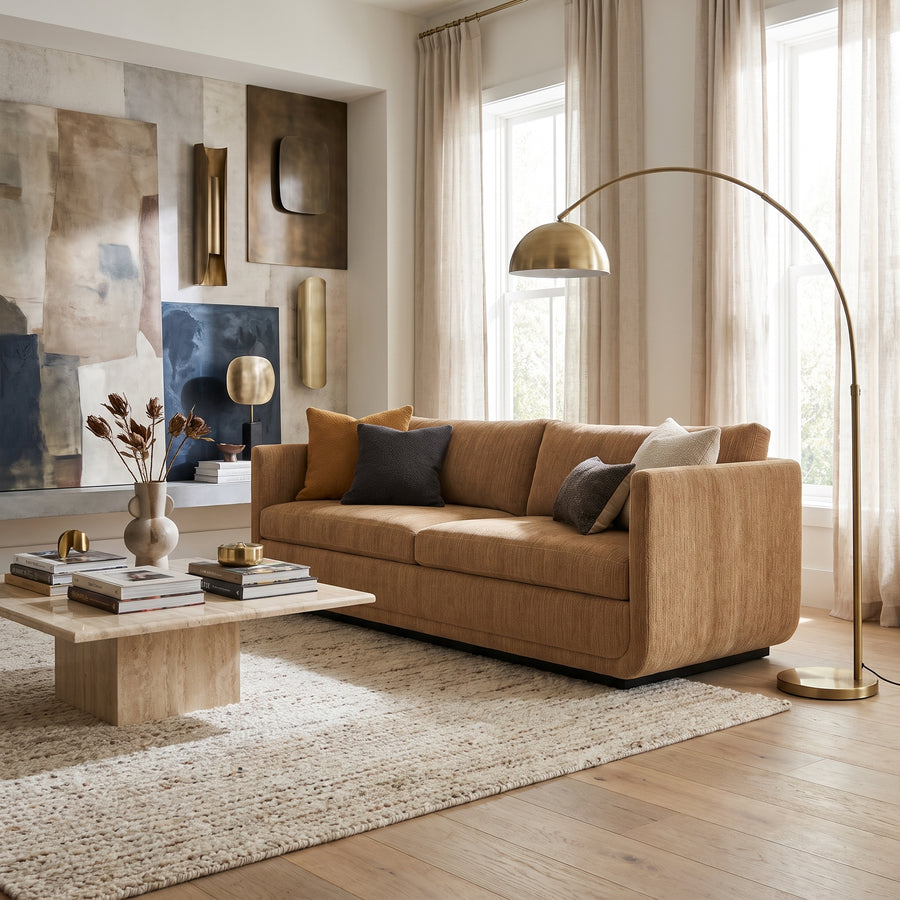 Modern living room with a tan sofa, travertine coffee table, arched floor lamp, and abstract wall art in a neutral palette.