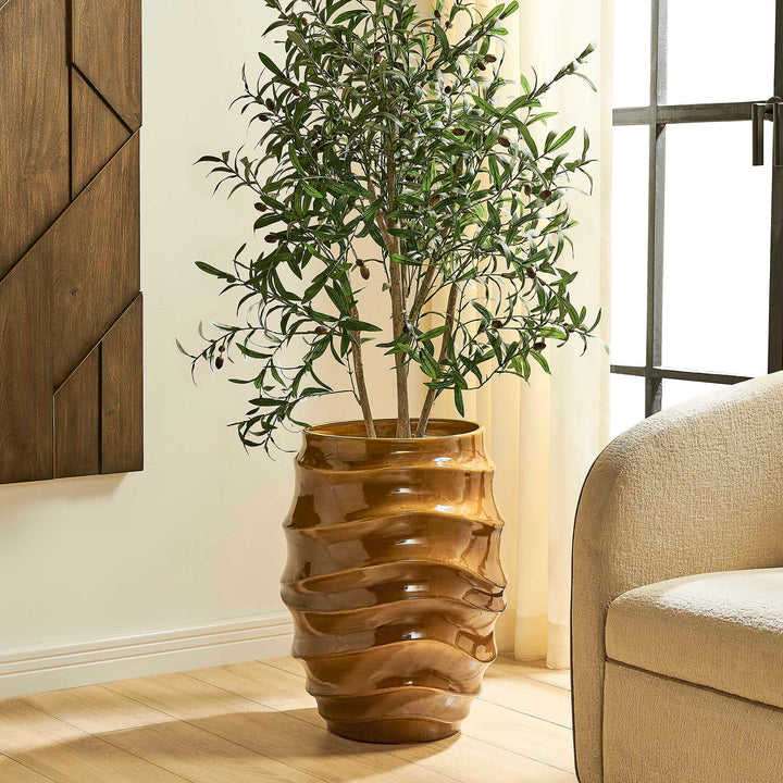 A potted olive tree with green leaves and small dark olives, displayed in a glossy, wavy ribbed brown ceramic pot on a light wood floor in a modern living room setting.