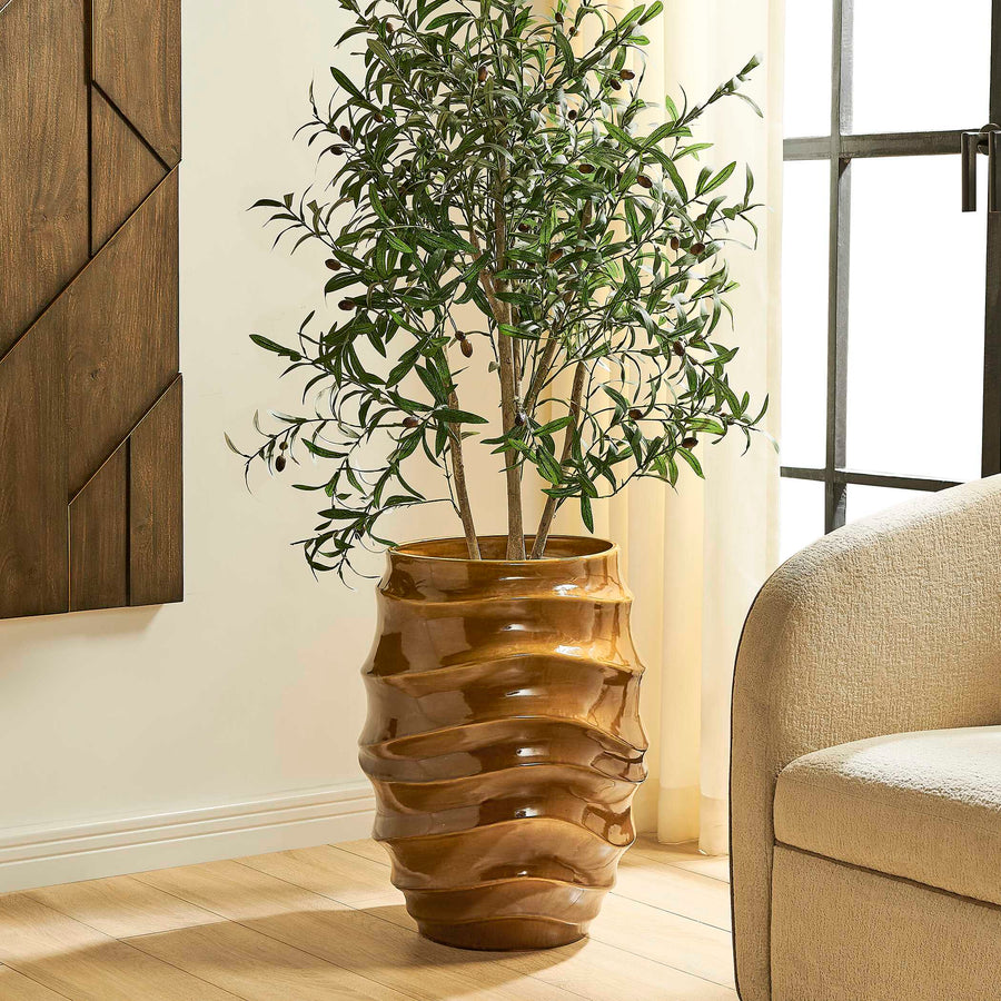 A potted olive tree with green leaves and small dark olives, displayed in a glossy, wavy ribbed brown ceramic pot on a light wood floor in a modern living room setting.