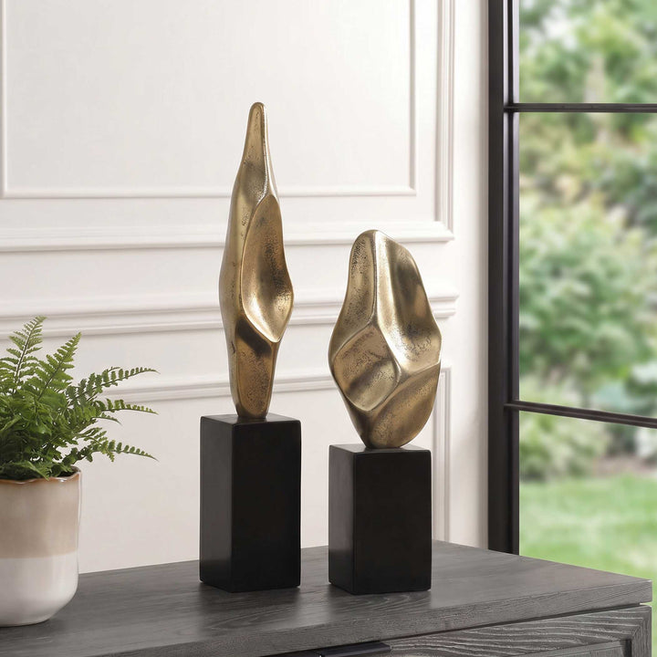 Two abstract metallic gold decorative sculptures on black rectangular pedestals, displayed on a dark wood console table in a styled interior setting with a potted fern and a window view.