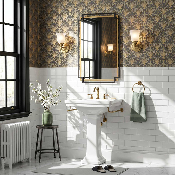 A styled bathroom interior featuring a white pedestal sink with brass fixtures, a gold-framed mirror, a green towel on a ring, and a decorative wallpaper pattern on the upper wall. A window, radiator, stool with flowers, and slippers are also visible.