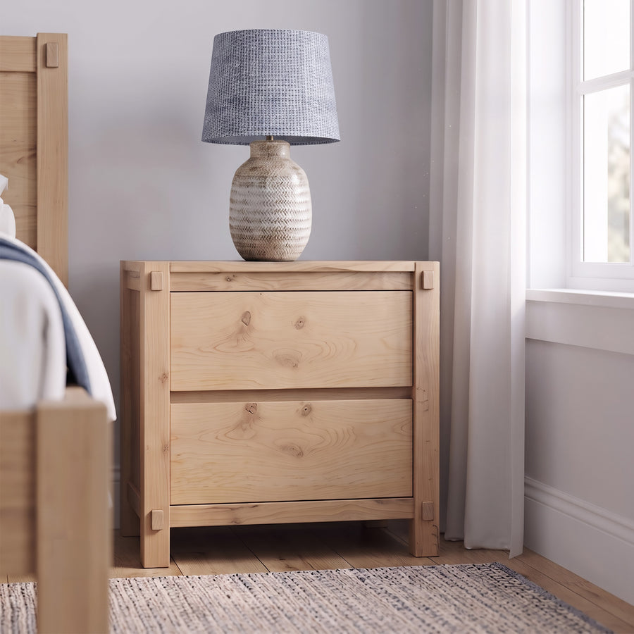 A light natural wood two-drawer nightstand in a bedroom setting, styled with a textured gray and beige table lamp on top.
