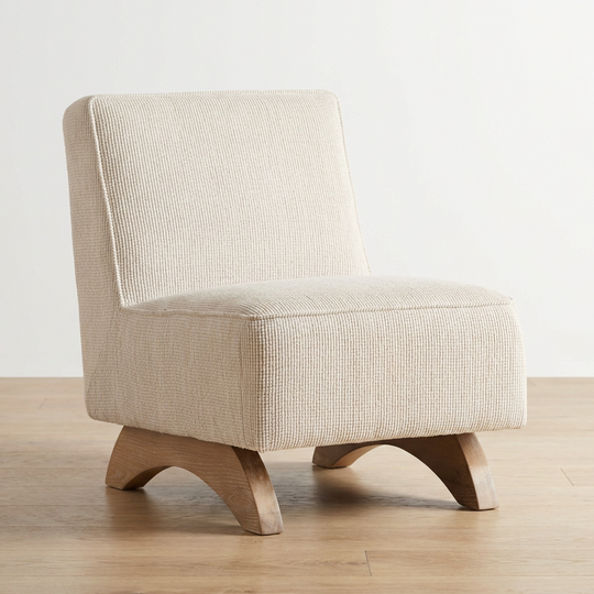 A modern low-profile accent chair upholstered in a textured beige fabric, featuring curved wooden legs, set against a minimalist white wall and light wood floor.
