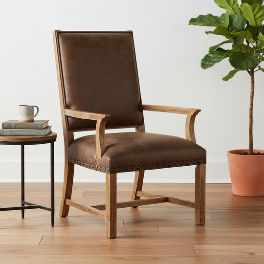 A brown leather upholstered armchair with a wooden frame and nailhead trim, shown in a styled room setting with a side table, mug, and potted plant.