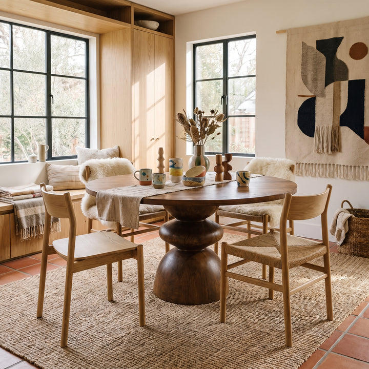 Modern dining room with a round wooden pedestal table, woven chairs, and natural, earthy decor in a bright, cozy space.