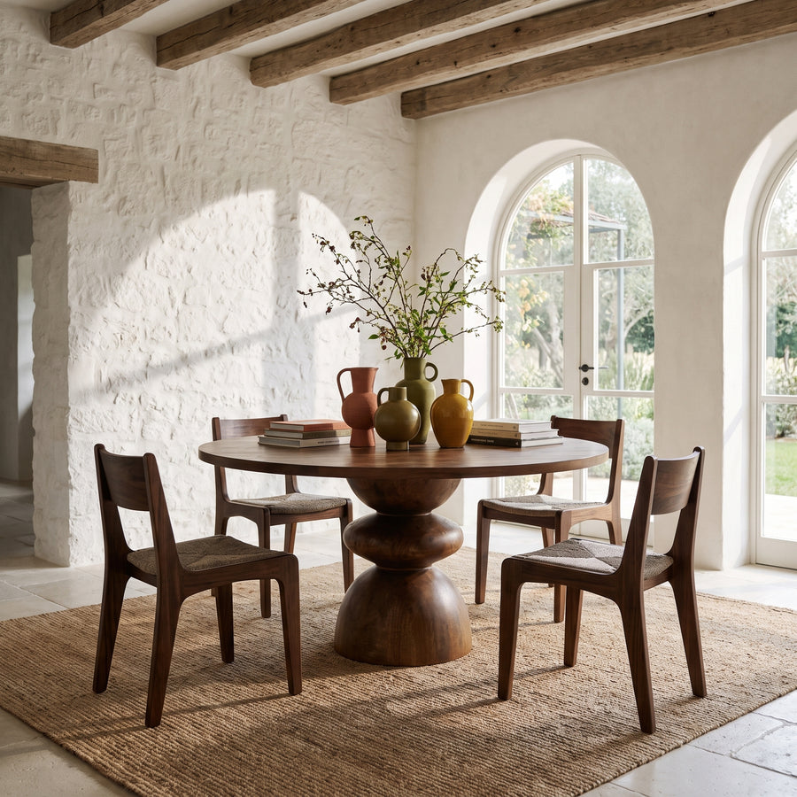 Rustic dining room with a round wooden pedestal table, modern chairs, and ceramic vases in a sunlit Mediterranean home.
