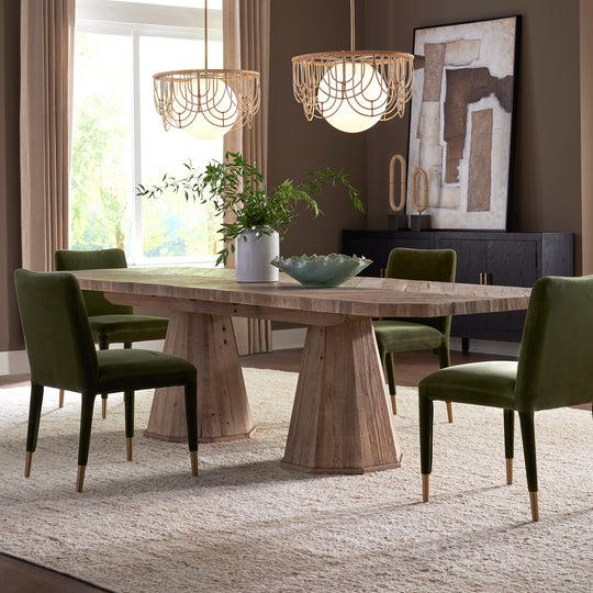 A reclaimed oak dining table with a herringbone inlay on a white background, accompanied by green chairs with gold accents. The room features a contemporary elegance with a geometric pedestal base.