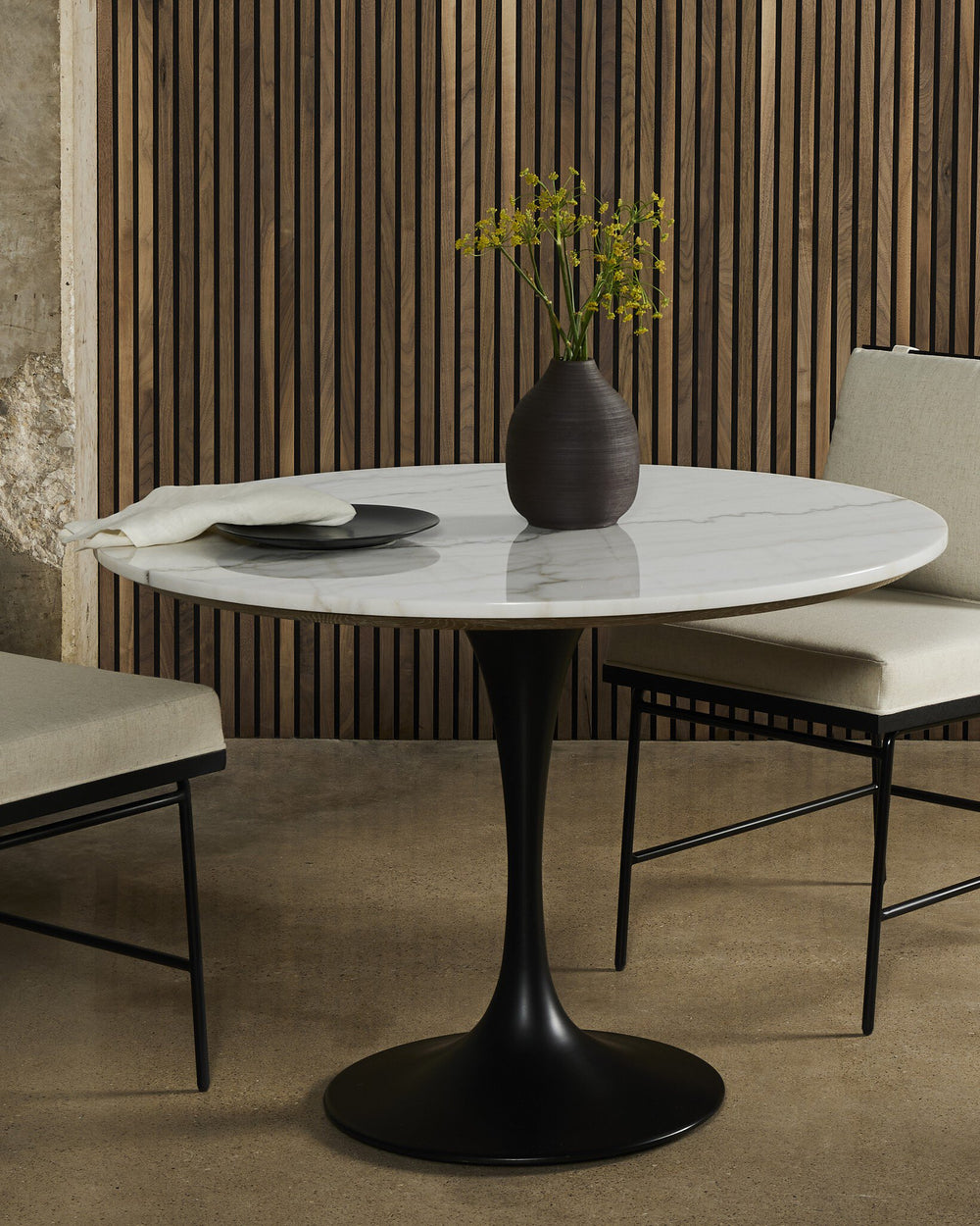 A modern dining table with a marble top, surrounded by two beige upholstered chairs, against a wood-slat wall and a vase of yellow flowers.