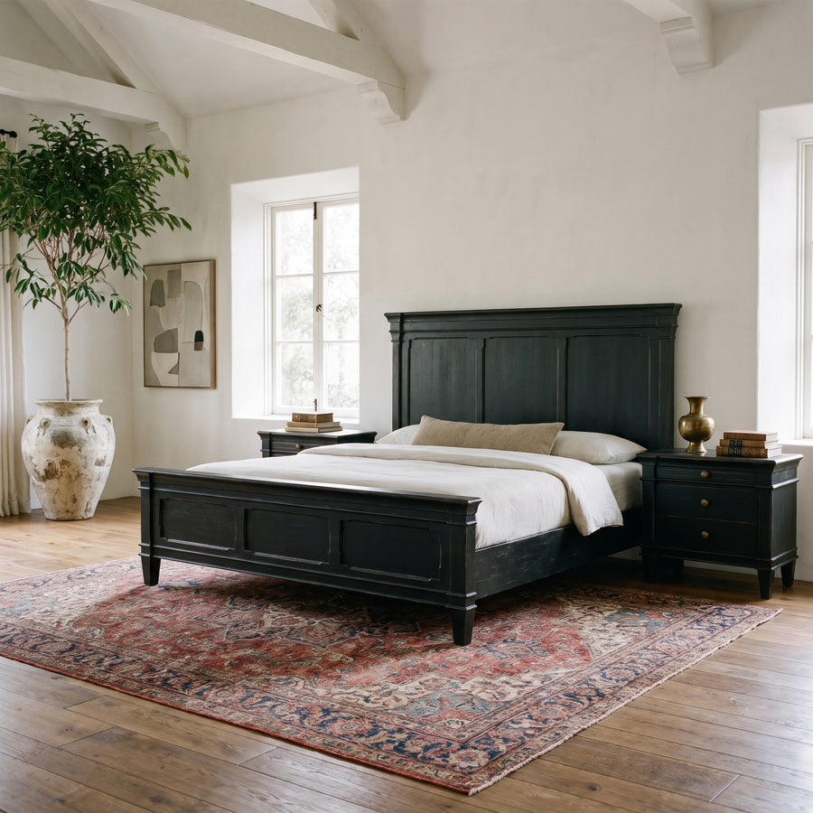 Rustic farmhouse bedroom with a black wooden bed frame, vintage area rug, and potted tree in a bright, airy space.