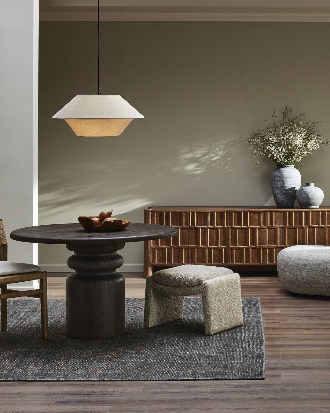 A modern dining area featuring a round table with a sculptural base, cozy seating, pendant lighting, and decorative vases.