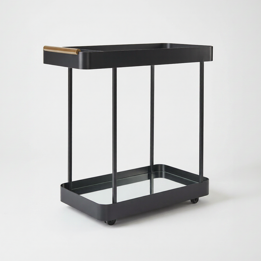 A modern two-tier bar cart in matte black metal with a clear glass lower tray and a gold top handle, shown on a plain light gray background.