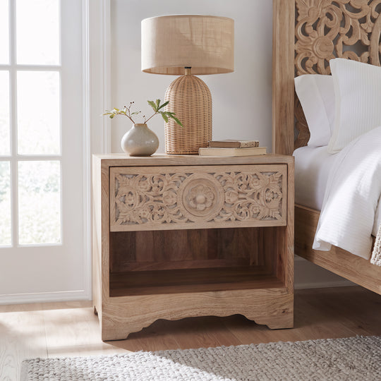 A light natural wood nightstand with ornate carved detailing on the drawer front