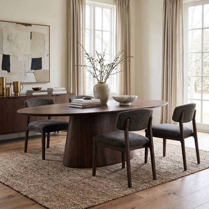 Modern dining room with a walnut oval table, upholstered chairs, and minimalist decor on a textured rug in a bright space.