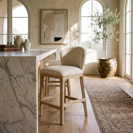 Modern farmhouse kitchen with a marble island, textured bar stools, an olive tree, and a vintage runner rug in a bright room.