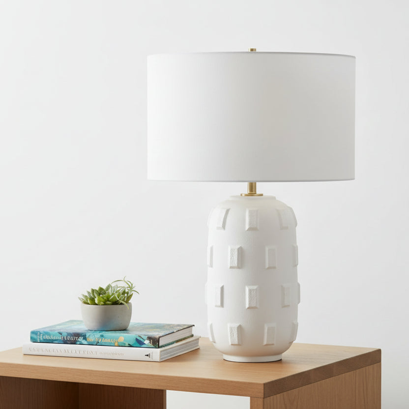 White textured table lamp on a wooden surface with books and a plant.