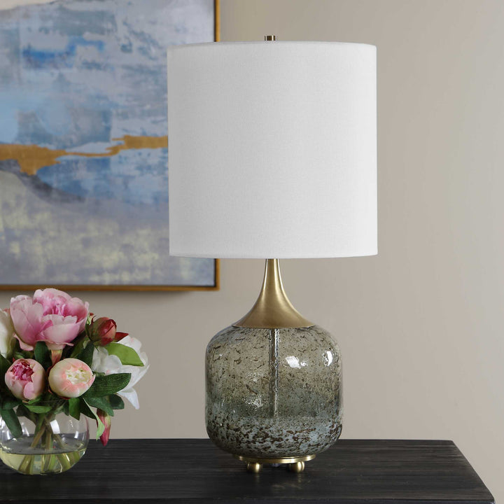 A table lamp with a white drum shade, a gold-finished neck, and a textured, speckled glass base in gray and green tones