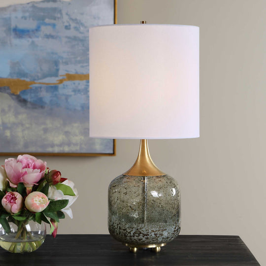 A table lamp with a white drum shade, a gold-finished neck, and a speckled glass base