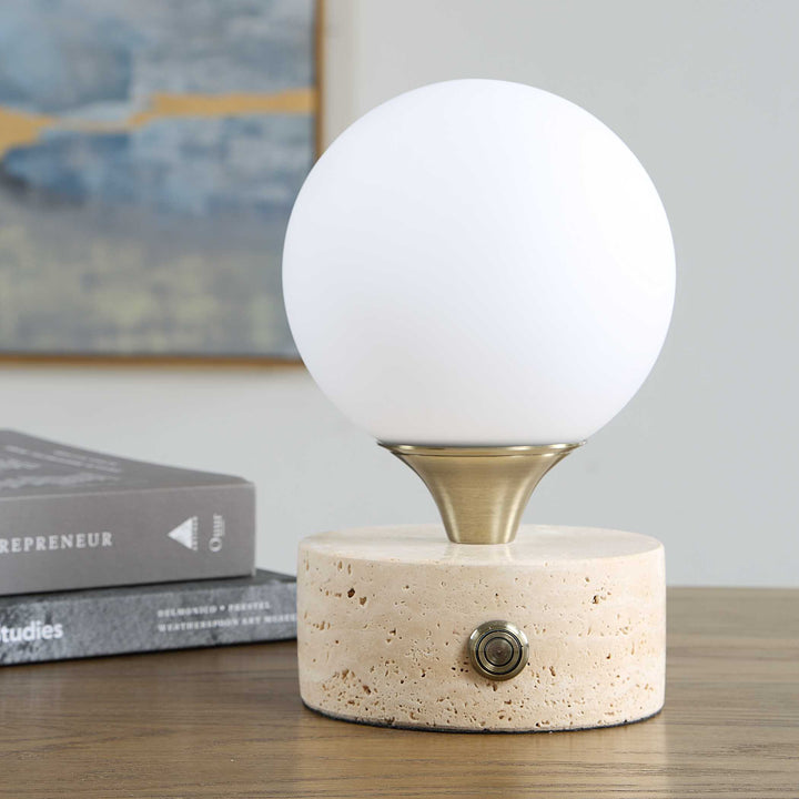 A modern table lamp featuring a white spherical globe on a brushed brass metal collar, mounted on a textured beige stone cylinder base with a single metal switch.
