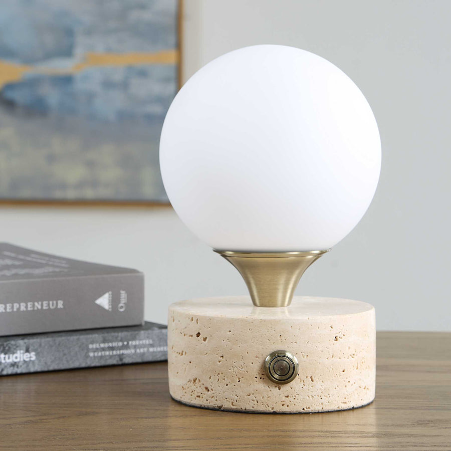 A modern table lamp featuring a white spherical globe on a brushed brass metal collar, mounted on a textured beige stone cylinder base with a single metal switch.