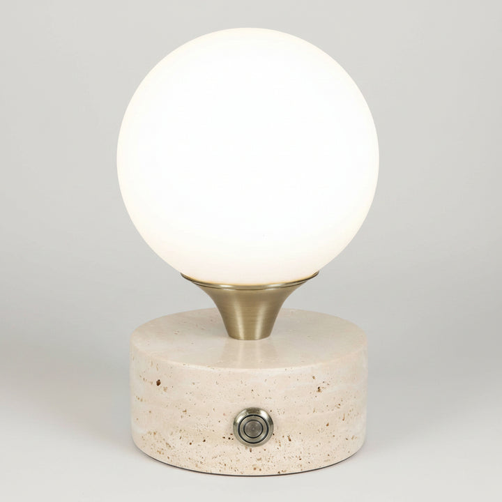 A modern table lamp featuring a glowing white spherical globe on a brushed brass metal collar, mounted on a textured beige stone cylinder base with a single metal switch.