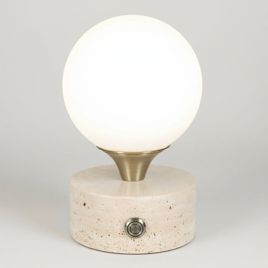 A modern table lamp featuring a glowing white spherical globe on a brushed brass metal collar, mounted on a textured beige stone cylinder base with a single metal switch.