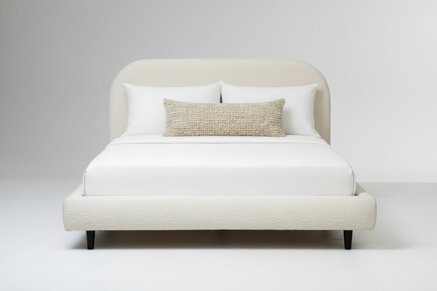 Beige upholstered bed with white bedding and a textured pillow on a plain background