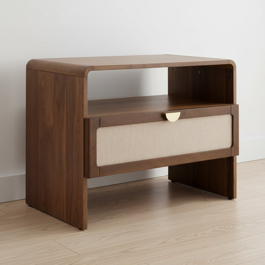 wooden nightstand, walnut finish, curved top edge, open shelf above drawer, beige fabric-front drawer with semi-circular brass handle, set against a white wall and light wood floor