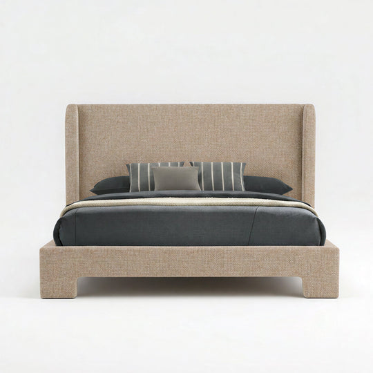 An upholstered bed frame with a tall wingback headboard and matching footboard, shown in a light beige fabric. The bed is styled with dark gray bedding, a striped gray pillow, and a solid gray accent pillow, set against a seamless white studio background.