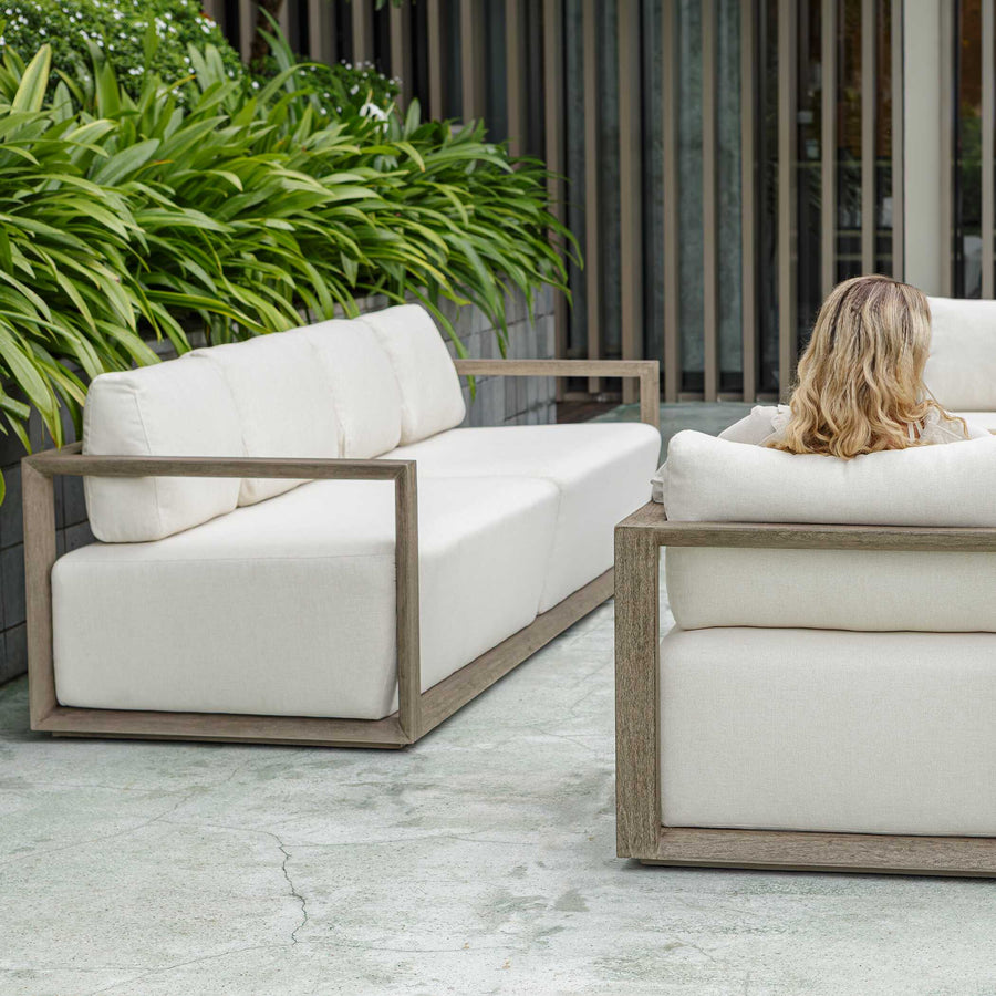 White outdoor sofa set with wooden frame on a patio with greenery and building in the background