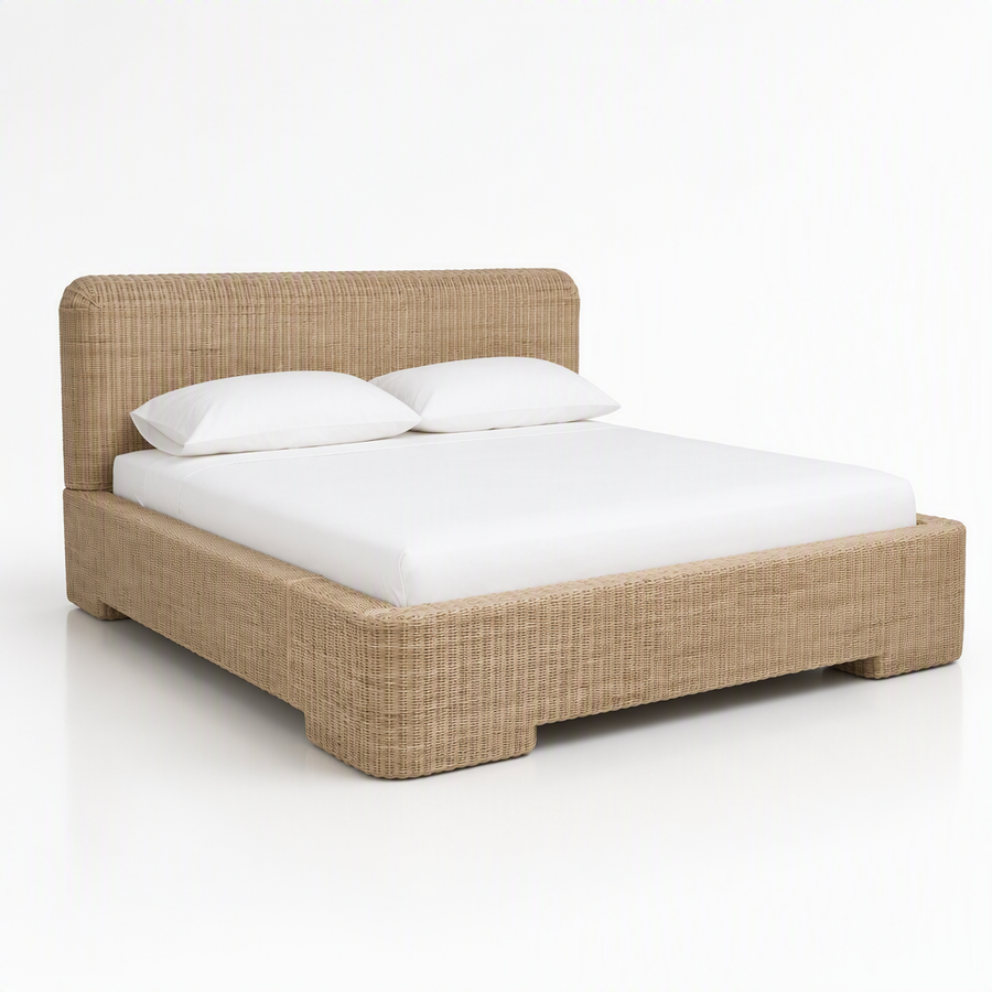 A low-profile platform bed with a woven beige rattan or wicker frame, dressed in a white mattress and two white pillows, set against a seamless white studio background.