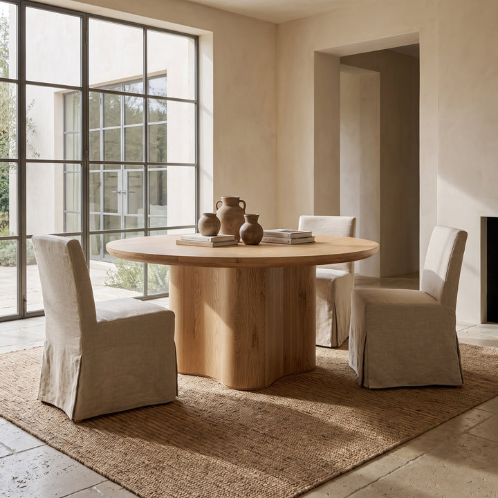 Modern minimalist dining room featuring a round oak pedestal table, linen slipcovered chairs, and a natural jute rug.