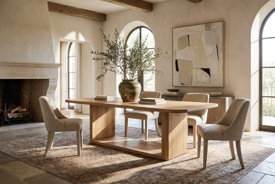 A modern dining room scene featuring a light oak wood dining table with a thick base and four upholstered dining chairs in a neutral beige tone, set on a patterned area rug in a sunlit room with a fireplace and arched windows.