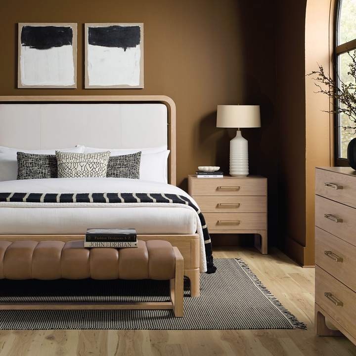 Wooden three-drawer nightstand with a natural finish, placed beside a bed.