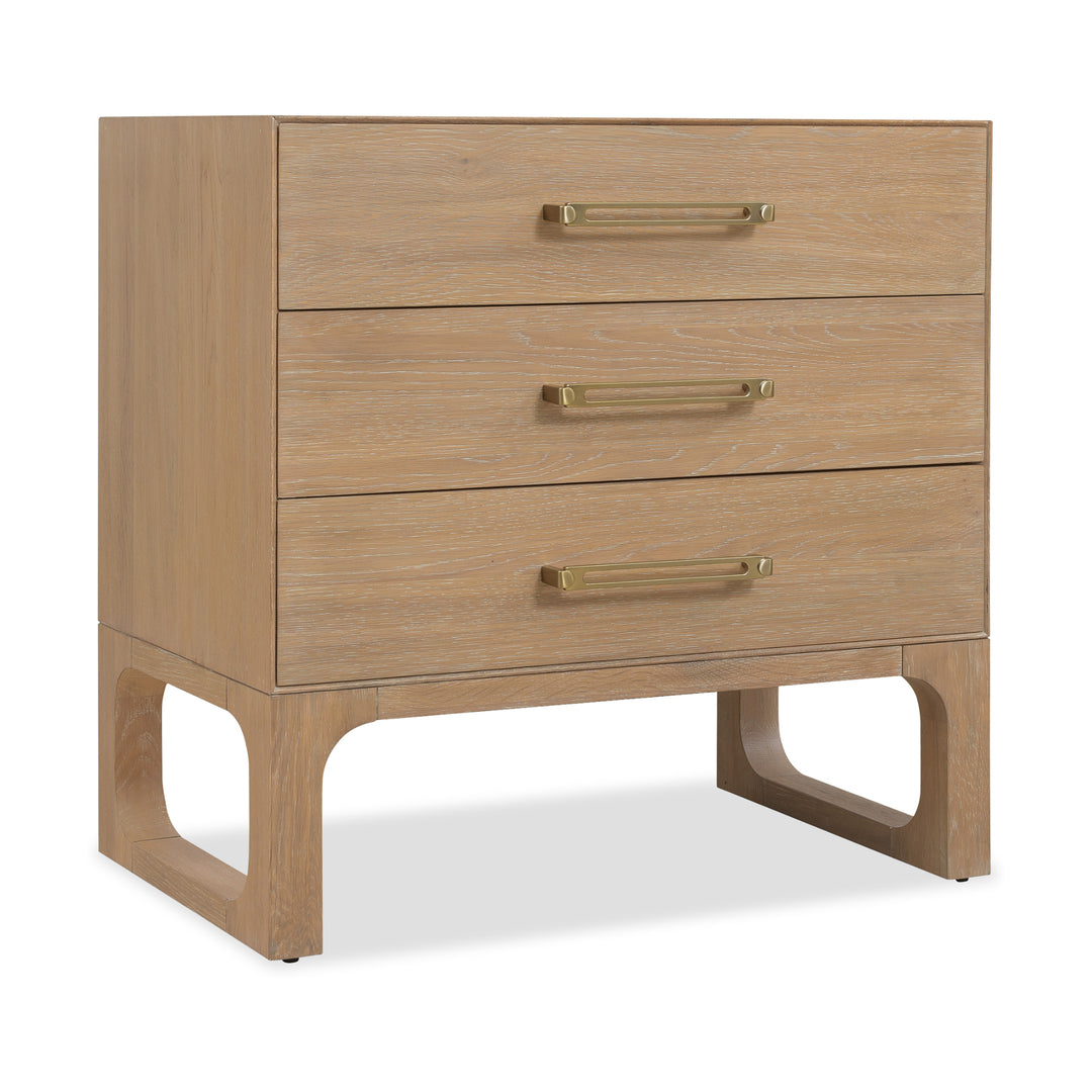 Angled view of a banyan wood nightstand with a light oak finish, three drawers, brass handles, and open-cut wooden legs for support.