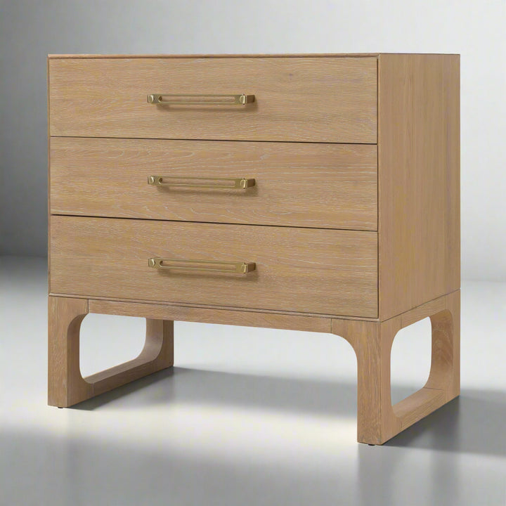 Angled view of a banyan wood dresser with a light oak finish, three drawers, brass handles, and open-cut wooden legs for support.