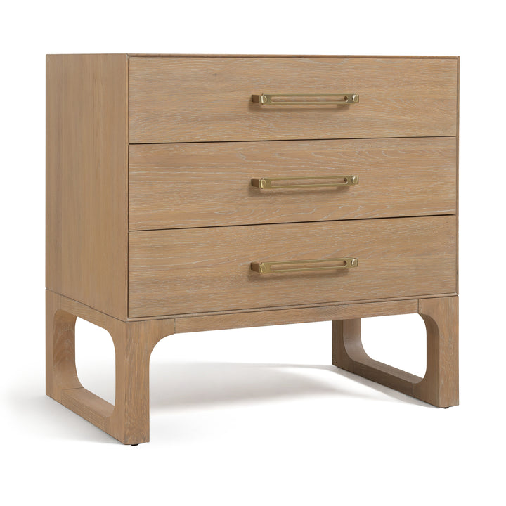 Angled view of a banyan wood dresser with a light oak finish, three drawers, brass handles, and open-cut wooden legs for support.