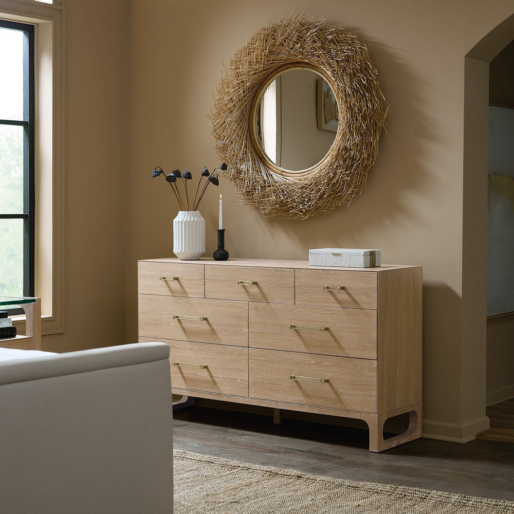 A modern banyon wooden dresser of 8 drawers topped with a vase, candle, and decorative box, beside a circular straw mirror on a soft beige wall.