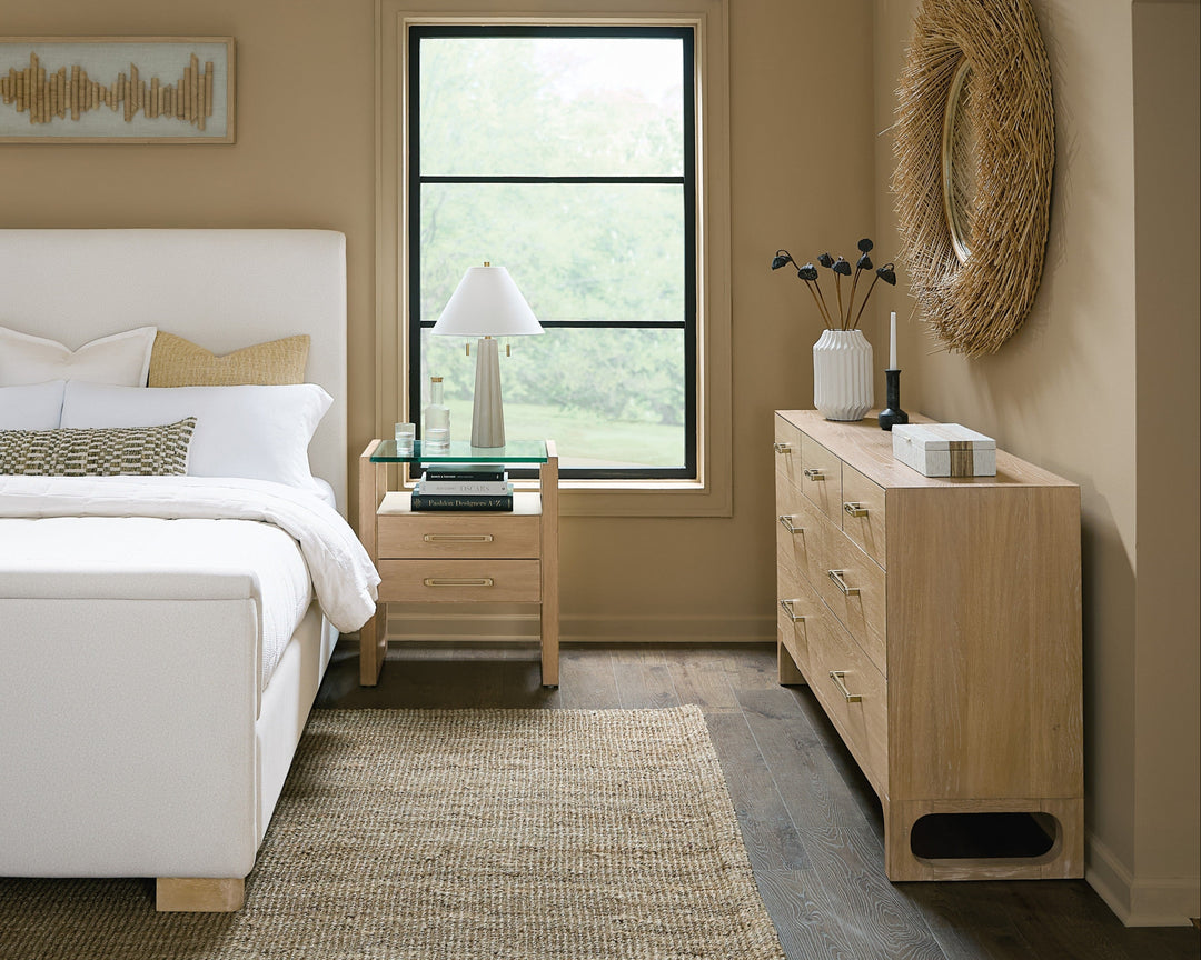 A modern bedroom featuring a white upholstered bed, wooden nightstand with a lamp, and a banyon wood dresser of 8 drawers against a beige wall.