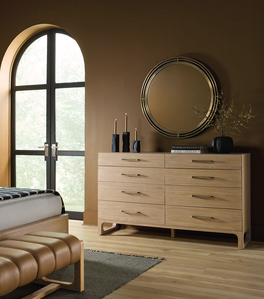 A sleek banyon wood dresser of 8 drawers with gold hardware, a round mirror, black candles, and a window, set against a rich brown wall.