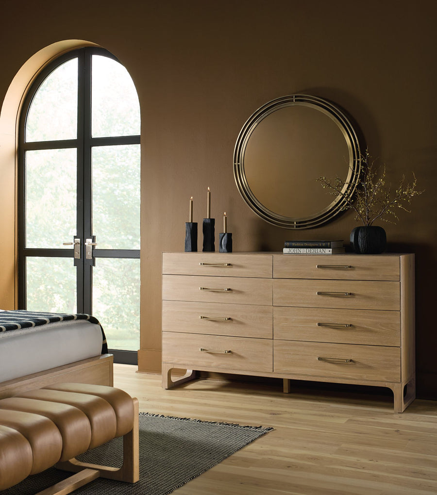 A sleek banyon wood dresser of 8 drawers with gold hardware, a round mirror, black candles, and a window, set against a rich brown wall.