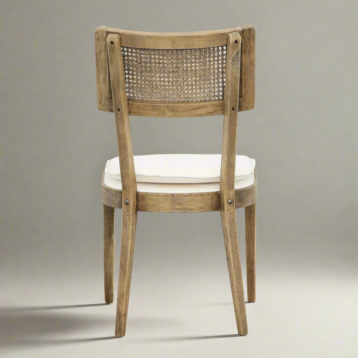 A wooden chair featuring a cane backrest and cream-colored cushion, viewed from the back against a neutral gray background.