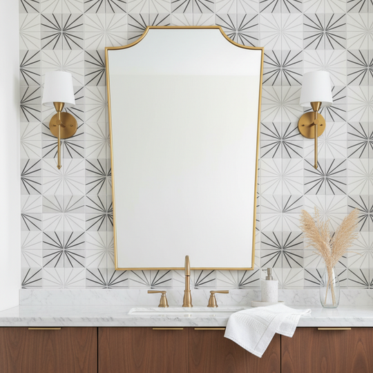 A large arched-top bathroom mirror with a gold frame hangs above a white marble sink and wooden vanity, flanked by two gold wall sconces against a patterned tile wall.