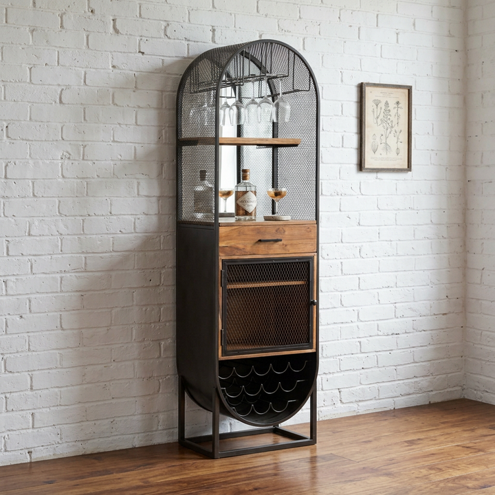 A tall industrial-style bar cart with a black metal frame, mesh wire shelves, and two wood-grain drawer fronts, shown against a white brick wall and wood floor.