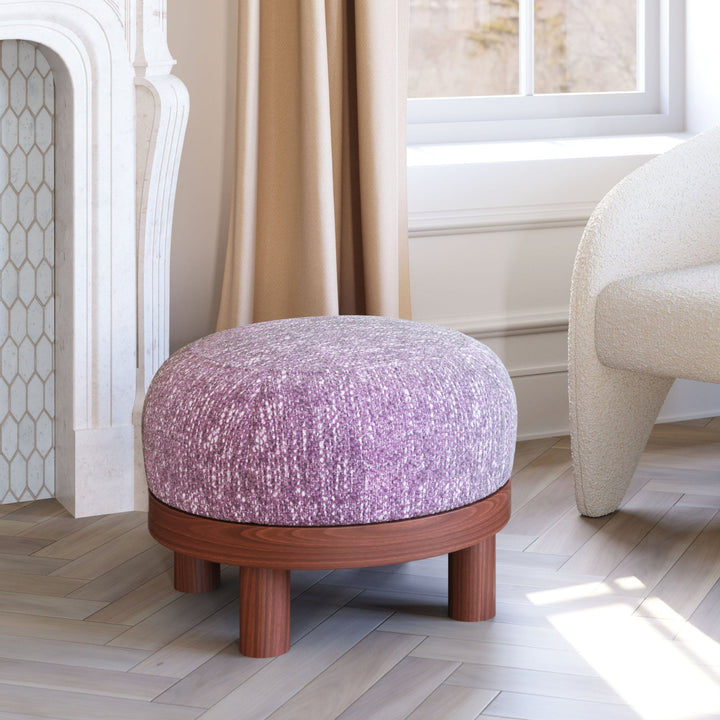 Purple ottoman with wooden base in a living room setting with a white armchair and fireplace.