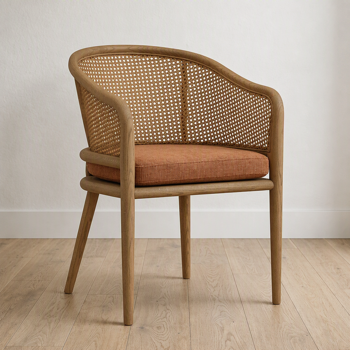 A wooden dining chair with a woven cane backrest and a rust-orange upholstered seat cushion, shown in a minimalist room setting with light wood flooring and a white wall.