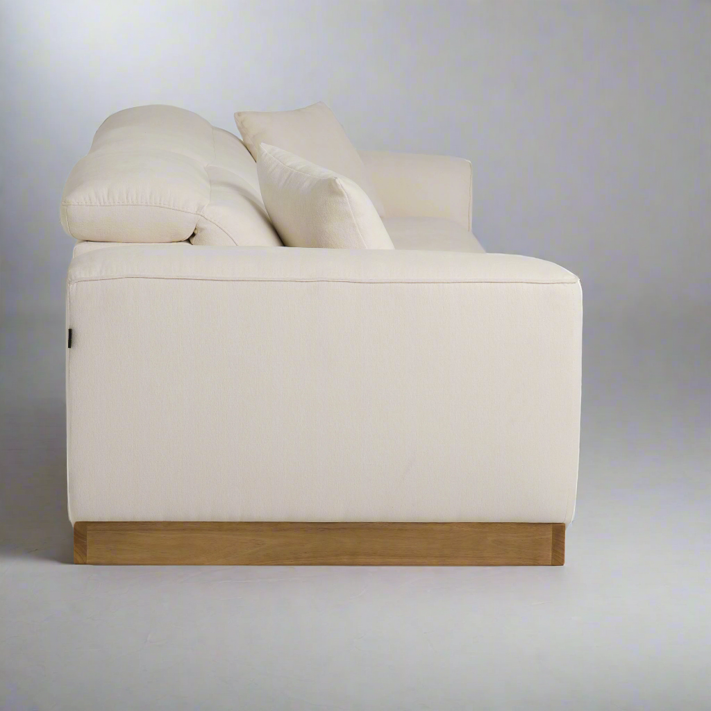 A modern white sofa with plush cushions and a wooden base, viewed from the side against a neutral gray background.